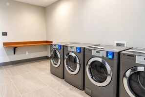 A laundry room with four front-loading washing machines, a wooden shelf, and a wall-mounted electrical outlet. - La Quinta Inn and Suites by Wyndham Central McKinney (McKinney)