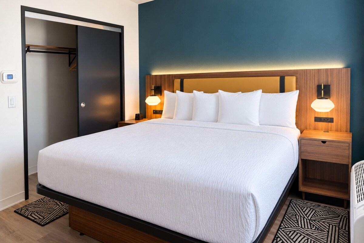A hotel room with a large bed, wooden headboard, two bedside tables with lamps, a wardrobe, and a patterned rug.