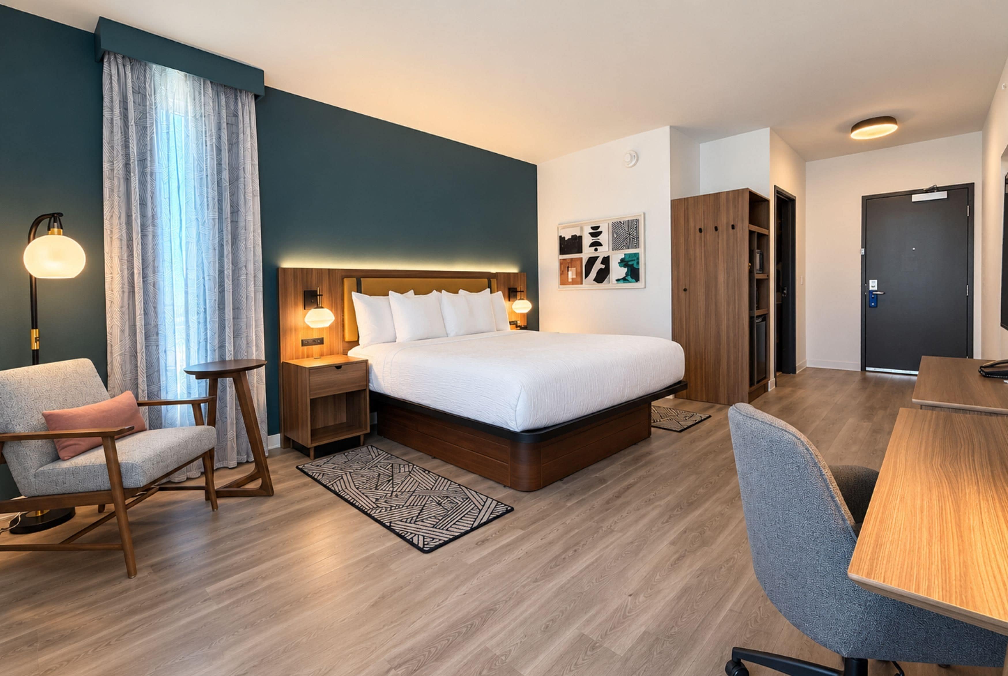 A modern hotel room with a large bed, bedside tables, a desk, a chair, and a wardrobe.