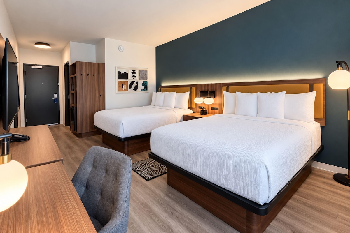 A hotel room with two beds, a desk, a chair, a wardrobe, and a wall with artwork.