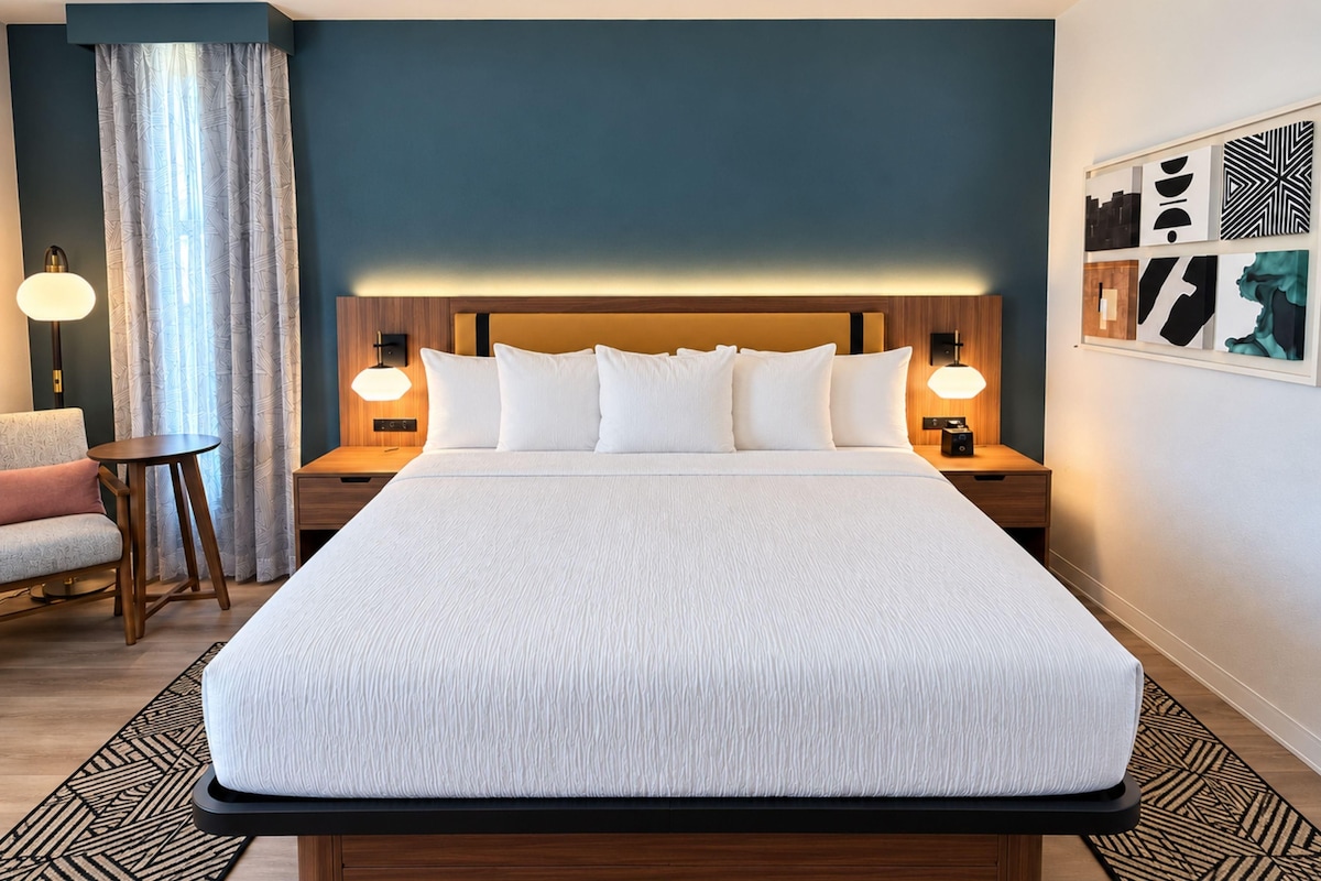 A hotel room with a large bed, two bedside tables with lamps, a small round table, a sofa, and a patterned rug.