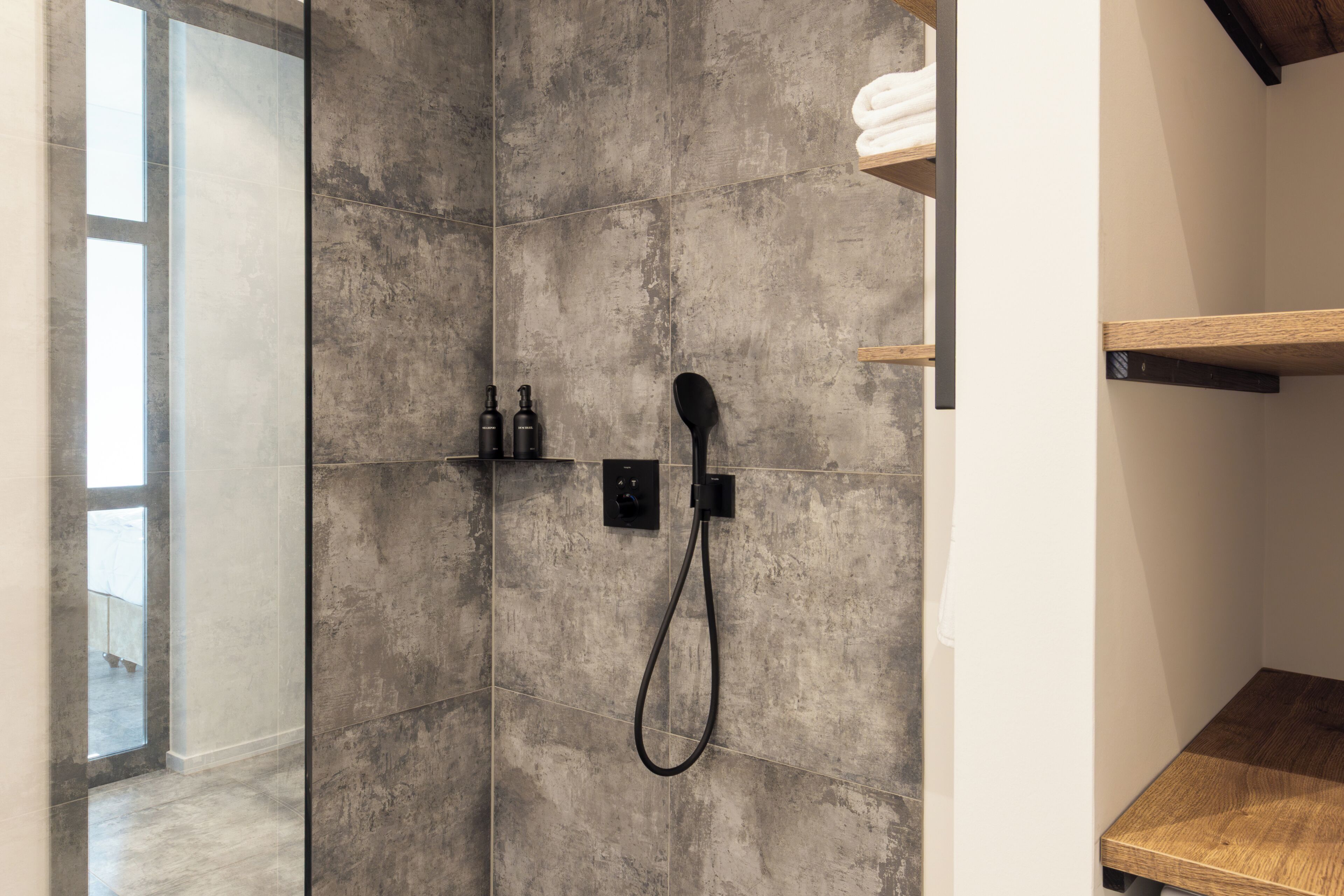 A shower area with a handheld showerhead, a wall-mounted control panel, and a shelf with toiletries.