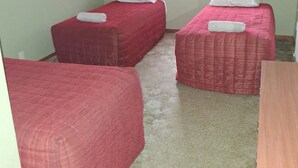 Habitación con dos camas, cada una con una colcha roja y un almohadón blanco. Hay una ventana con cortinas, y las paredes están pintadas de un color claro.
