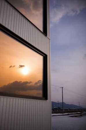 Une fenêtre donnant sur un coucher de soleil au-dessus d’une étendue d’eau, avec une ligne électrique et des montagnes lointaines en arrière-plan.