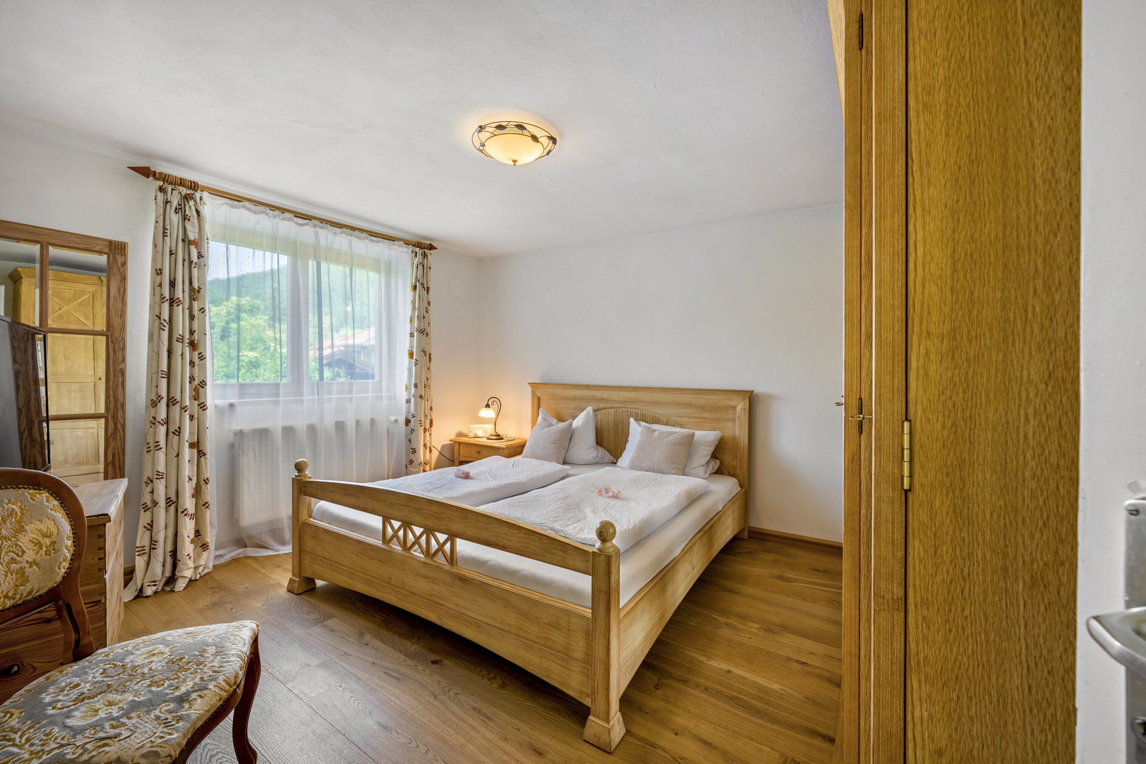 Ein Bett aus Holz in einem Raum mit Holzboden, einem Stuhl, einem Nachttisch mit Lampe und einem Fenster mit Vorhängen.