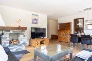 A living room with a stone fireplace, a flat-screen TV, a wooden cabinet, and a dining area with chairs and a table. - Panoramic view, 2 bedrooms, covered parking (Nendaz)