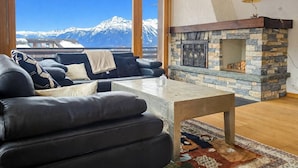 Ein Wohnzimmer mit Kamin aus Stein, einer Sofagarnitur und Blick auf schneebedeckte Berge.