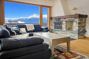 A living room with a stone fireplace, a sofa set, and a view of snow-capped mountains. - Panoramic view, 2 bedrooms, covered parking (Nendaz)