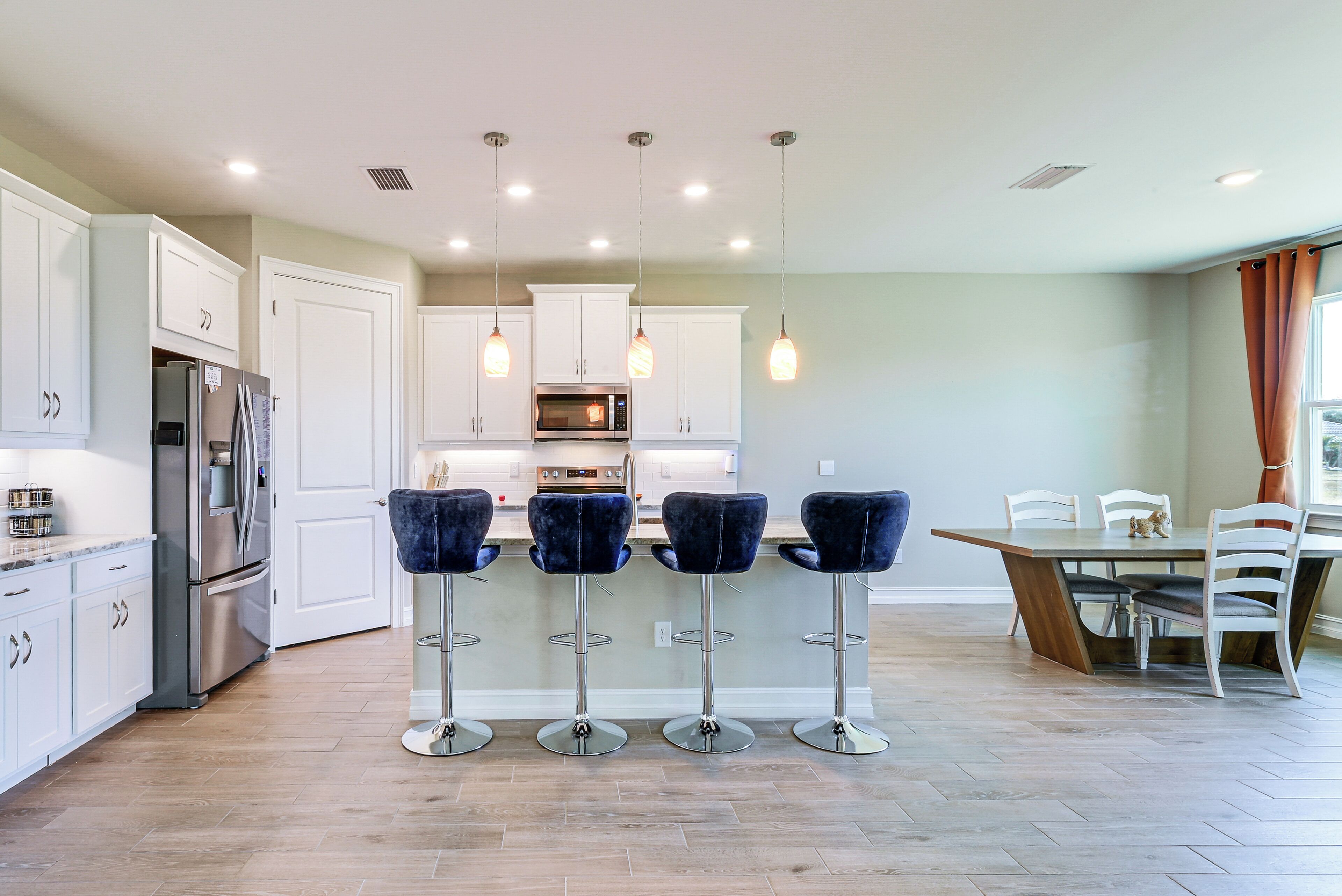 Una cocina moderna con armarios blancos, un refrigerador de acero inoxidable, un microondas y un comedor con una mesa y sillas de madera.