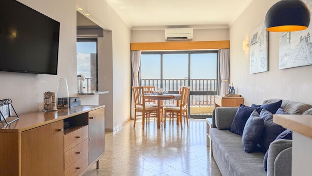A modern living room with a flat-screen TV, a wooden dining table with chairs, a grey sofa, and a balcony with a view. - #357 Sea View Getaway in Portimão | Pool & Spacious (Portimão)