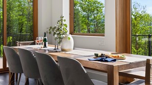 Un coin repas avec une table en bois, des chaises, un vase avec une plante et une vue sur les arbres à travers de grandes fenêtres.