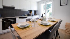 A modern kitchen with white cabinets, a wooden dining table set for four, and a view of the outdoors through a large window. - Stunning, bright apartment with a private garden (Farvagny-le-Grand)