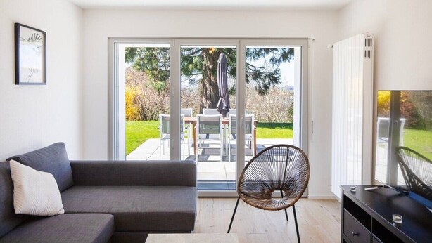 A modern living room with a sofa, a chair, a TV stand, and a sliding glass door leading to an outdoor patio with a table and chairs. - Stunning, bright apartment with a private garden (Farvagny-le-Grand)