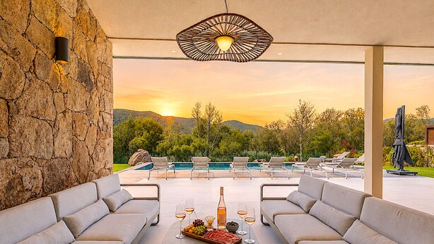 A poolside area with a stone wall, two sofas, a table with a bottle and glasses, and a view of the sunset over a pool and mountains. - Villa Déoni (Zonza)