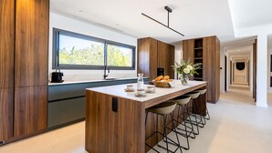 A modern kitchen with a central island, bar stools, a large window, and wooden cabinetry. - Villa Déoni (Zonza)