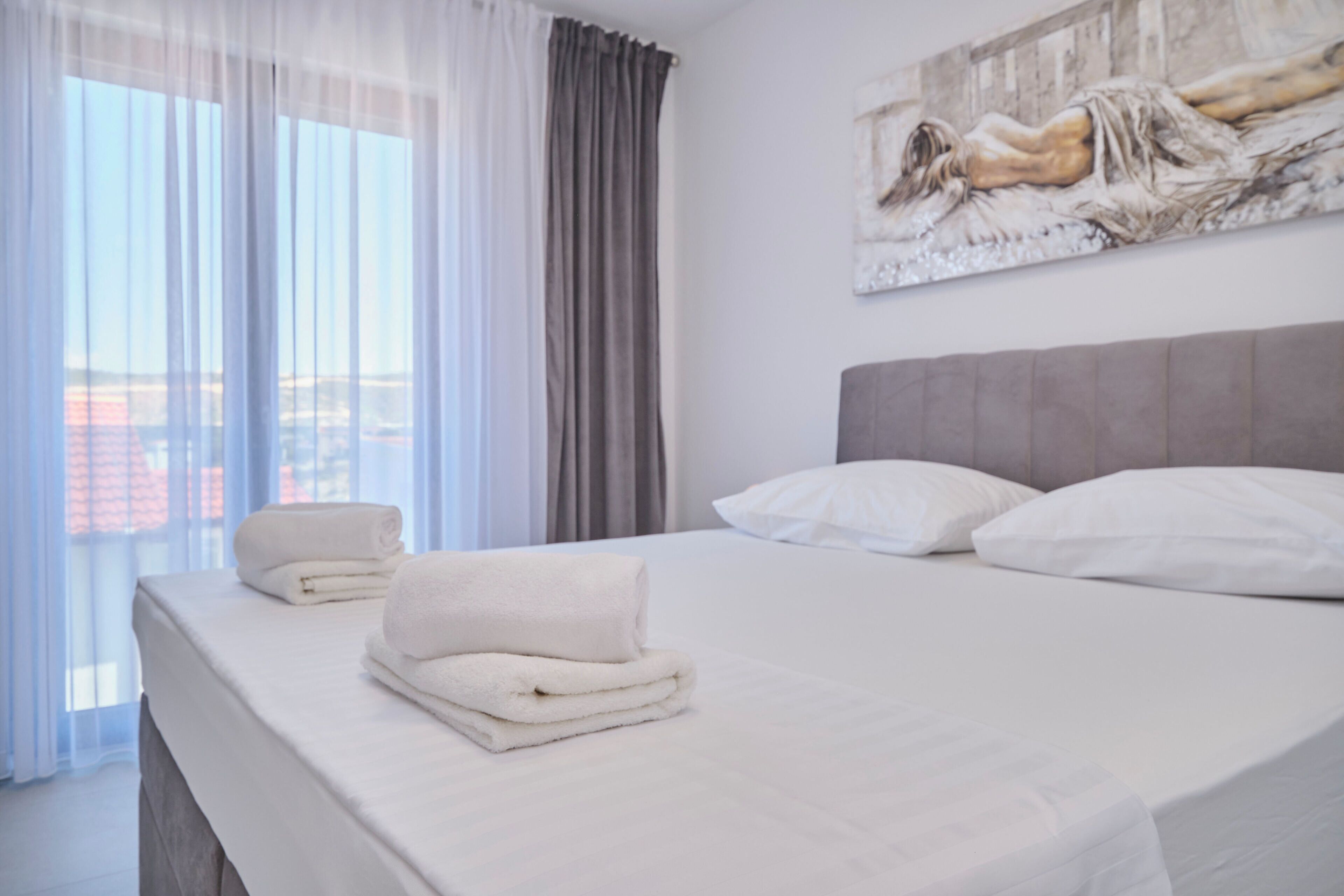 A hotel room with a bed, pillows, a painting on the wall, and a window with curtains.