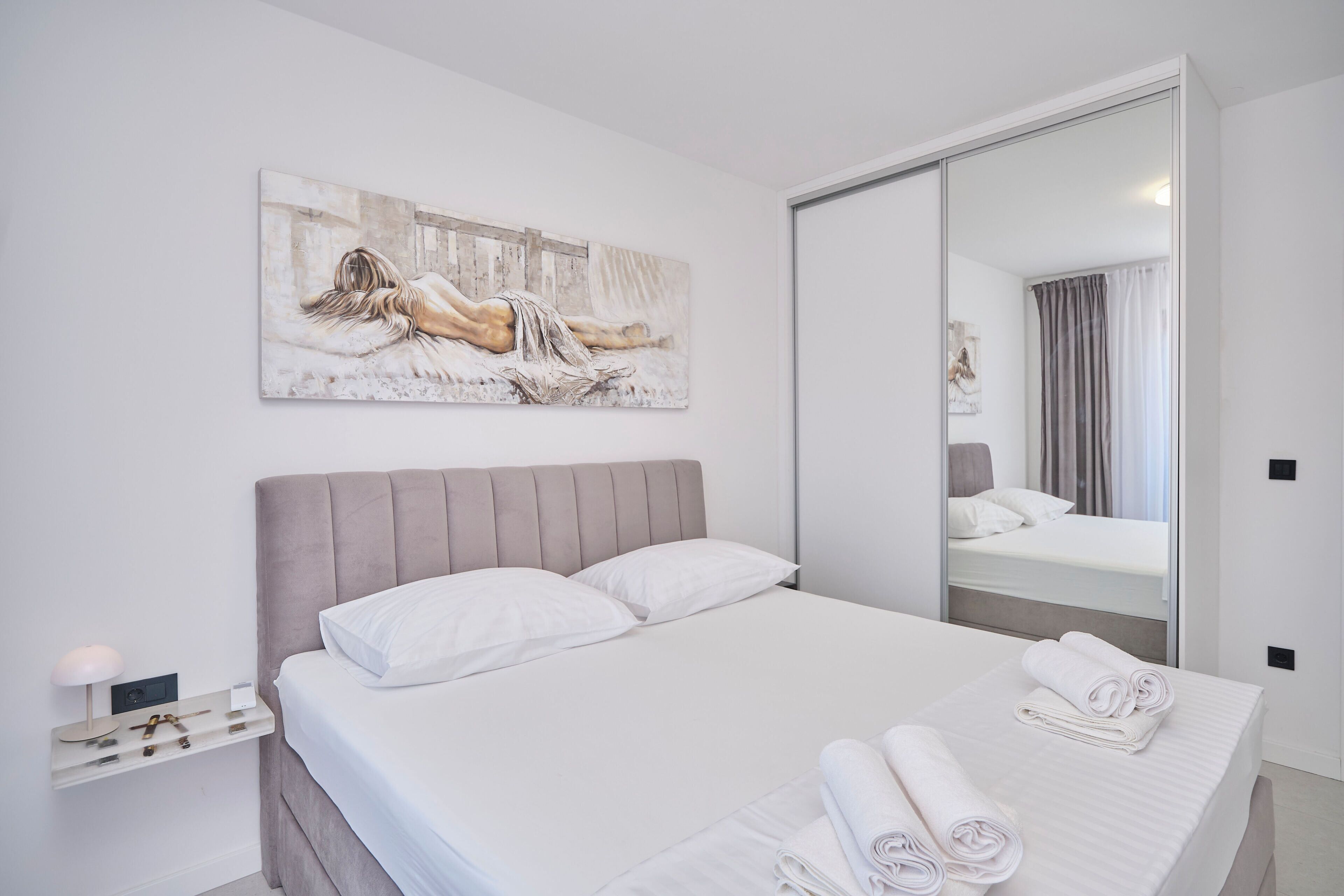 A modern bedroom with a large bed, a bedside table, a mirror, and a painting on the wall.