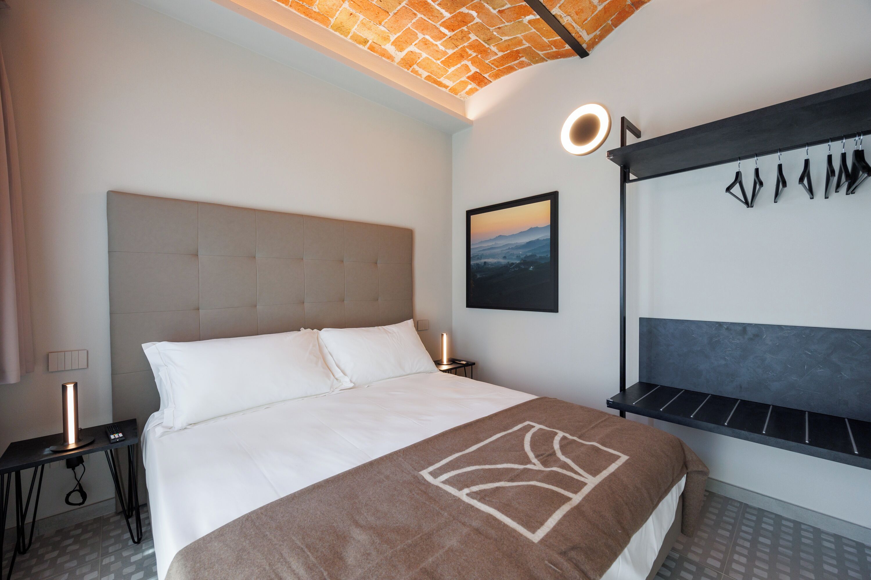 A modern hotel room with a large bed, a headboard, a bedside table, a nightstand, a wall-mounted light, a framed picture, and a unique wall art piece.