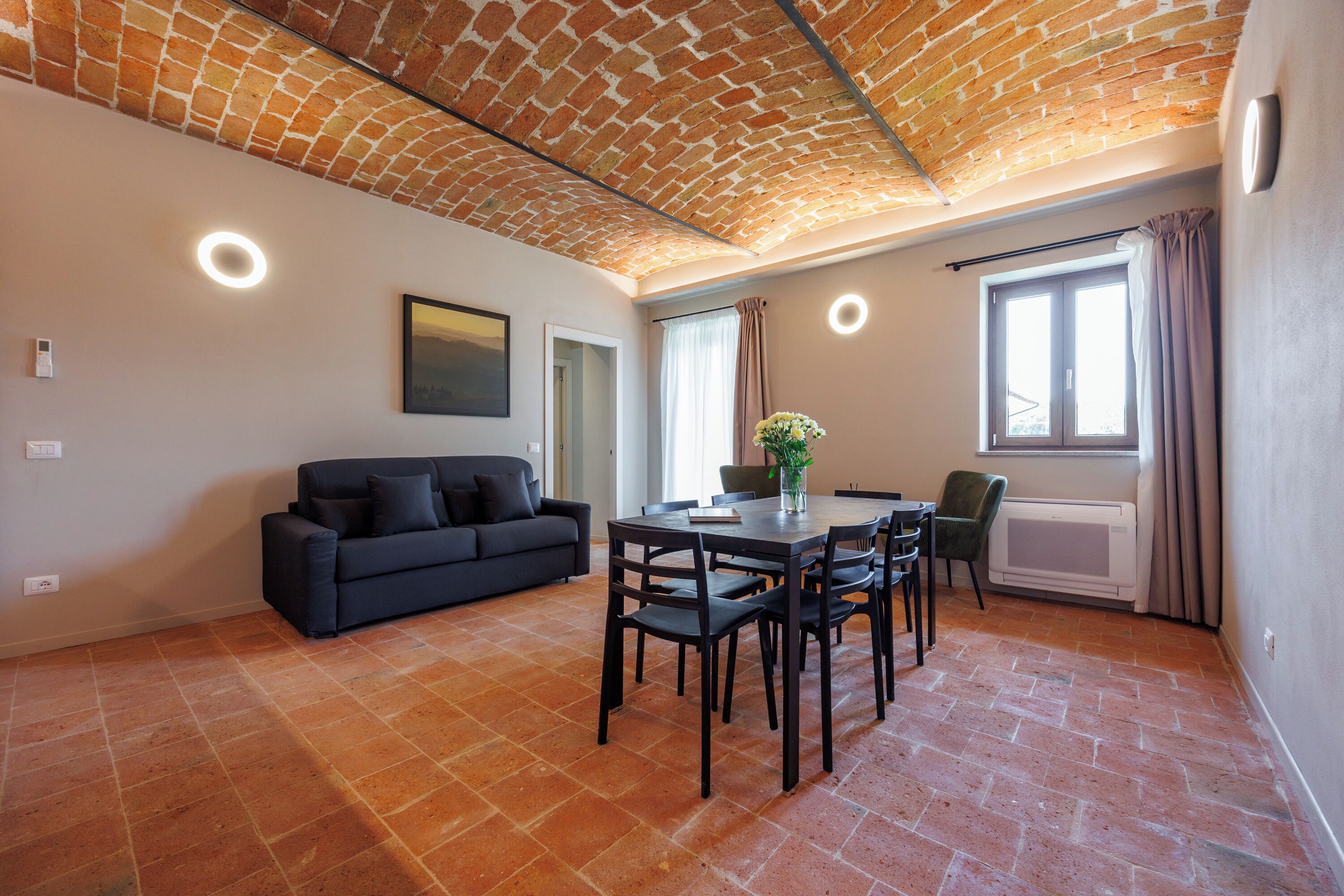Una sala de estar moderna con techo de ladrillo, un sofá negro, una mesa de comedor con sillas y un ventanal con cortinas.