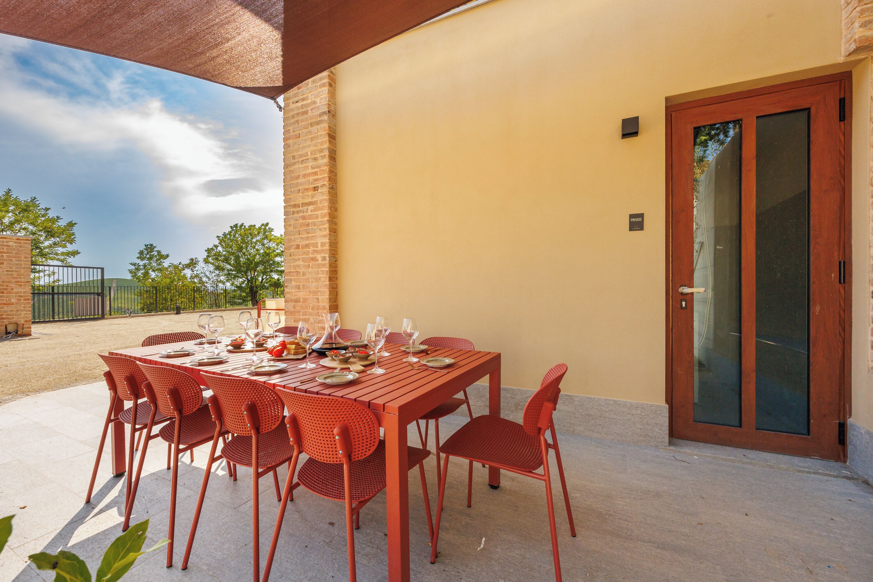 A covered outdoor dining area with a brick wall, a sliding glass door, and a table set for six with red chairs.