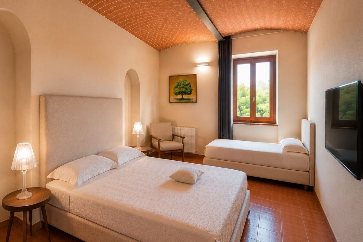 A hotel room with a large bed, a smaller sofa, a chair, a TV, and a window with a view.