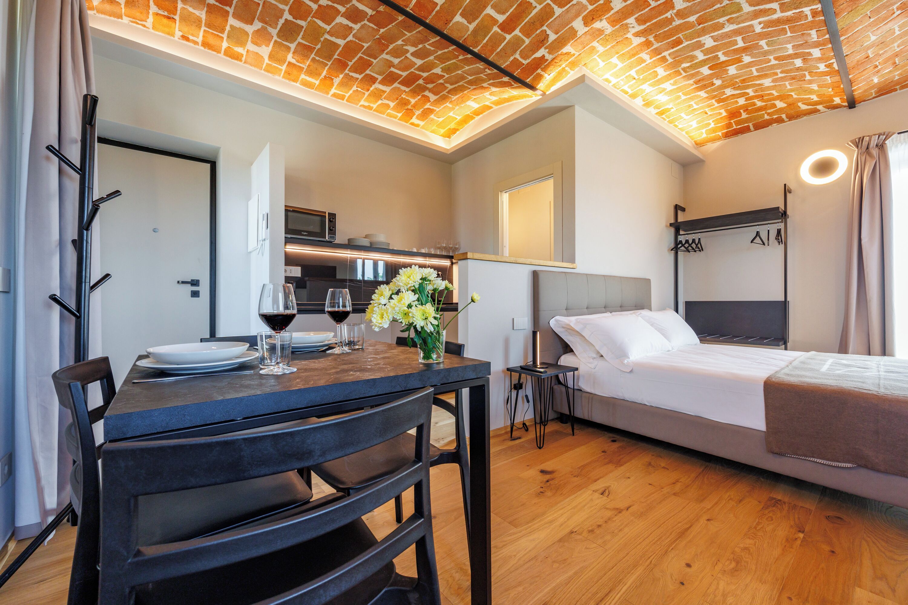 Una habitación moderna con techo de diseño ladrillo, una mesa de comedor con sillas, una cama con cabecera y una mesita con un jarrón de flores.