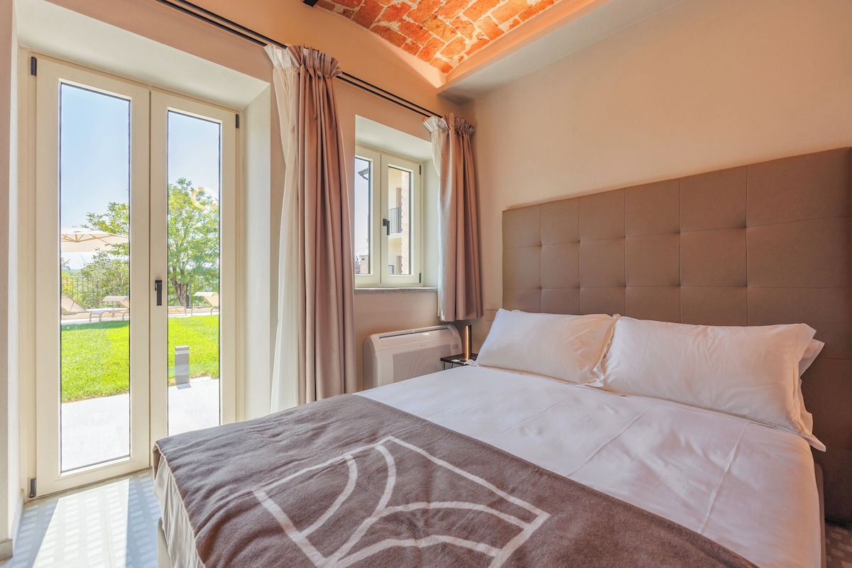 A bedroom with a large bed, a window with curtains, and a view of a green outdoor area.