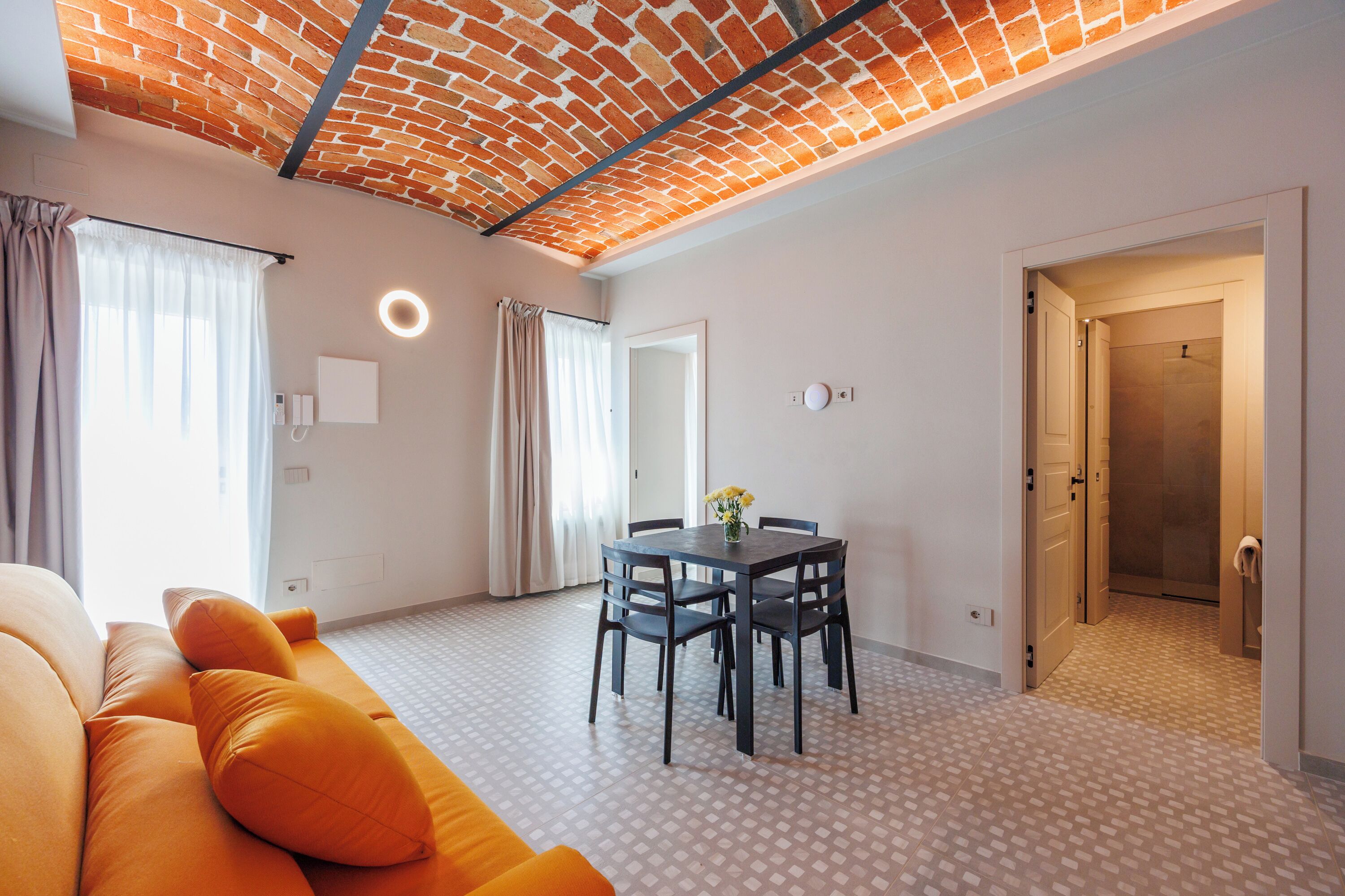 A modern living room with a brick-patterned ceiling, a dining area with a table and chairs, and a visible bathroom door.