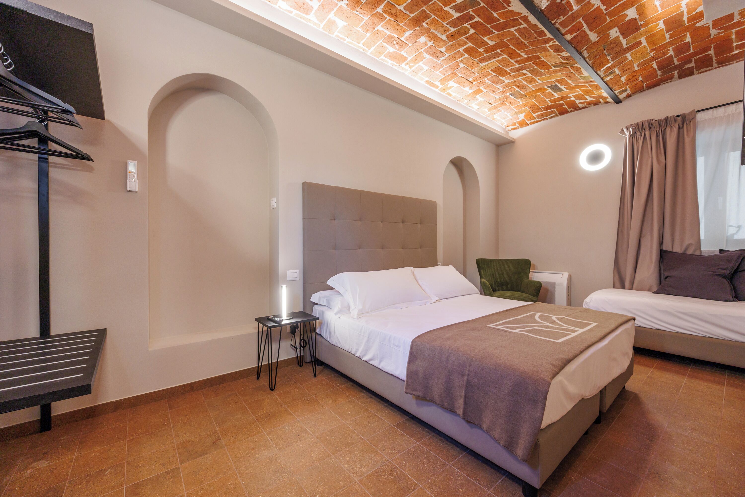A hotel room with a bed, a bedside table with a lamp, a chair, and a second bed.
