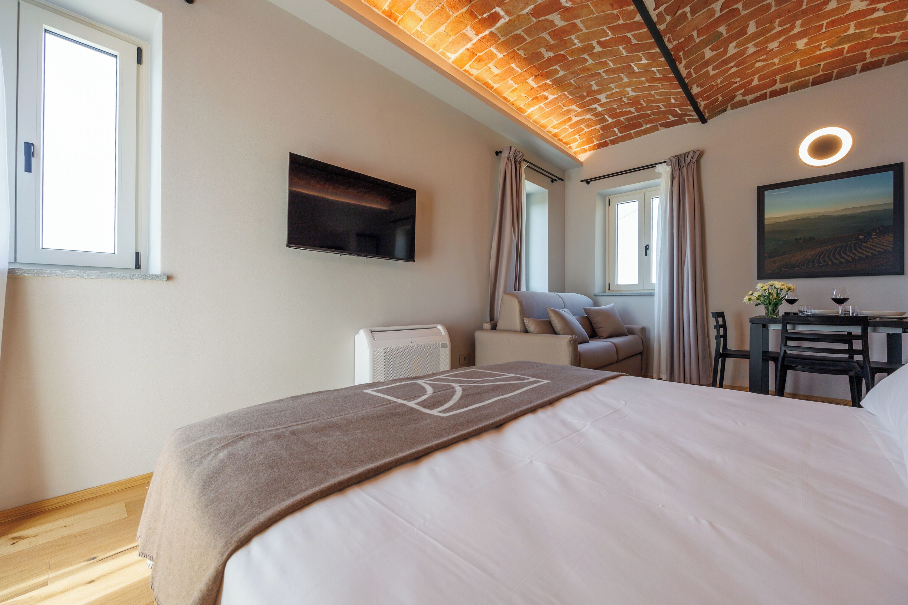 A hotel room with a bed, a flat-screen TV, a heater, a dining table with chairs, and a painting on the wall.