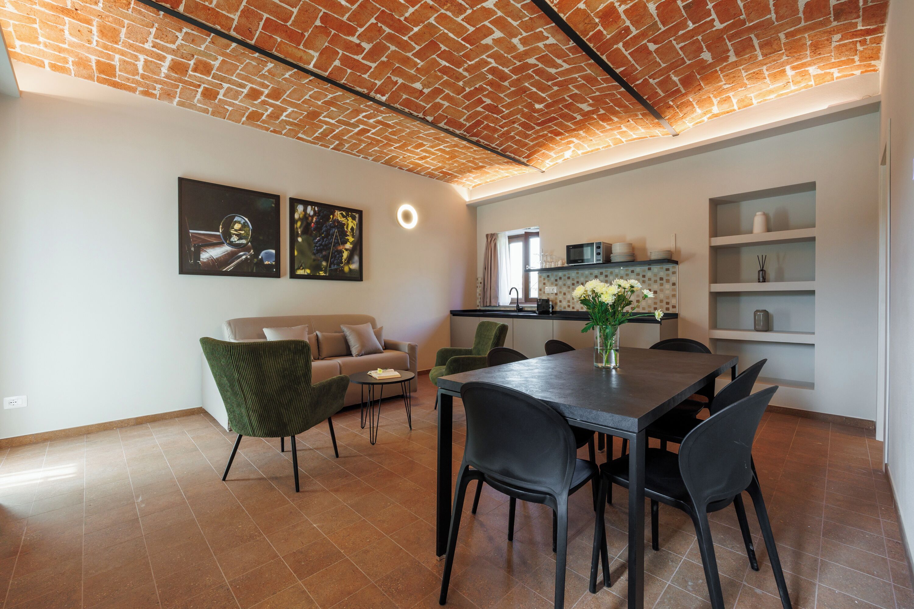 Um interior moderno com teto de tijolos, uma sala de jantar com mesa e cadeiras, uma cozinha com micro-ondas e prateleiras, e duas fotos emolduradas na parede.