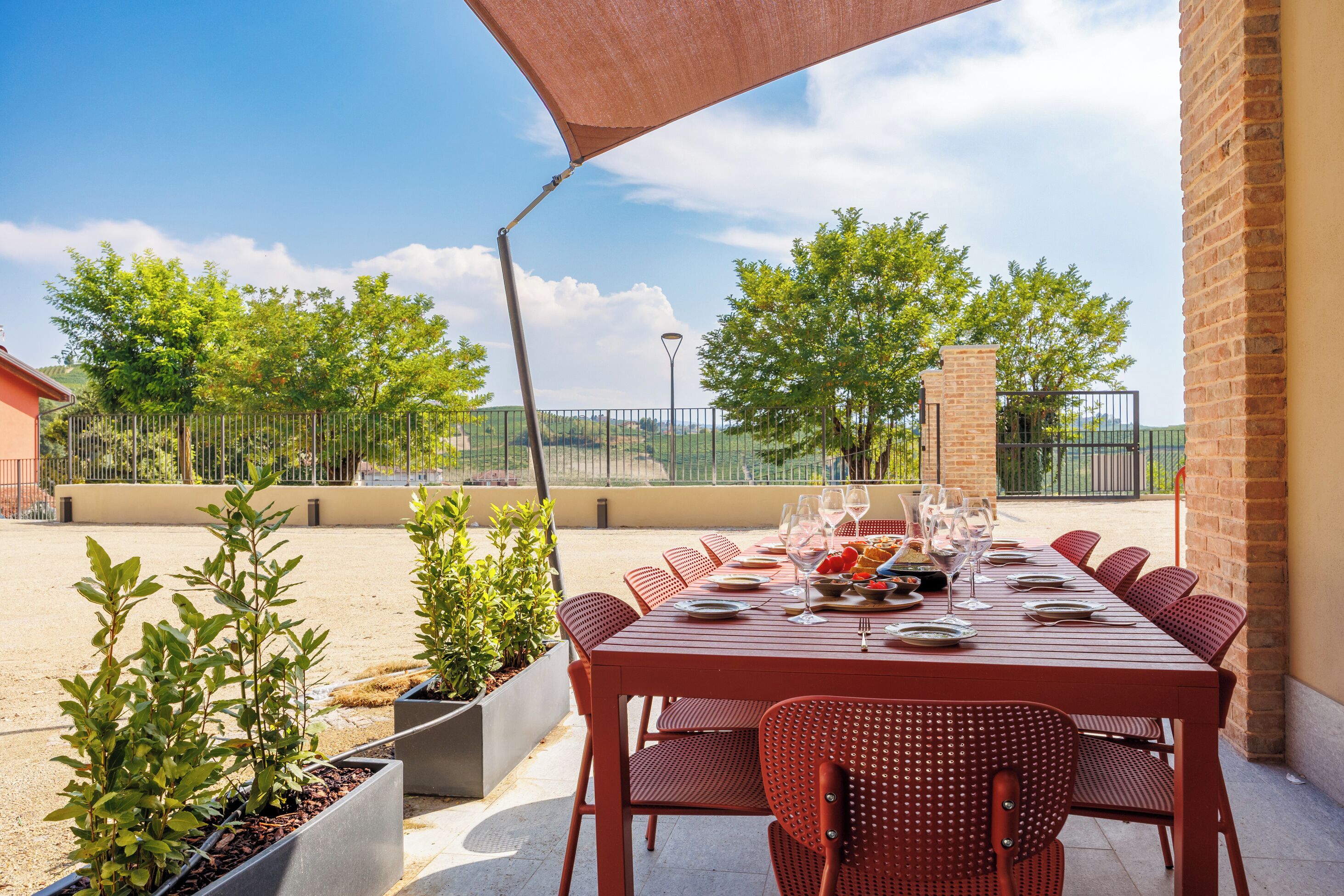 Un área de comedor al aire libre con una mesa preparada para seis personas, rodeada de sillas rojas, bajo un gran paraguas.
