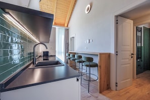 A modern kitchen with a black countertop, a stainless steel sink, and a wooden ceiling. There are bar stools and a door leading to another room. - Calanga (Barbaresco)