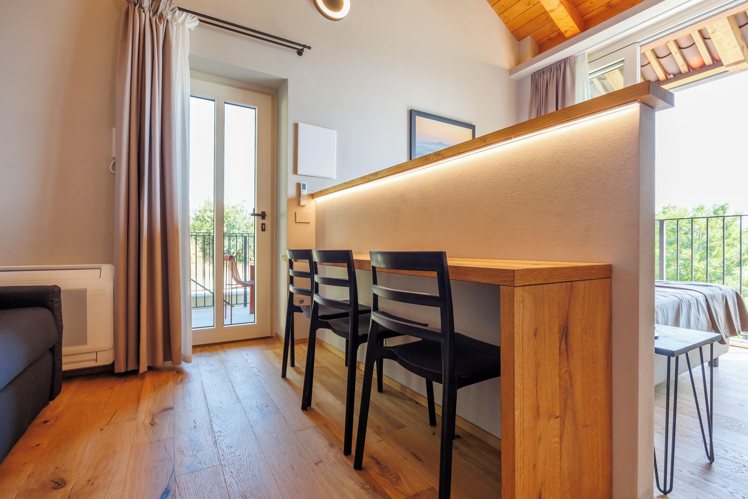 A compact living space with a kitchenette, a dining area with chairs, a sliding glass door leading to an outdoor balcony, and a bed with a view.