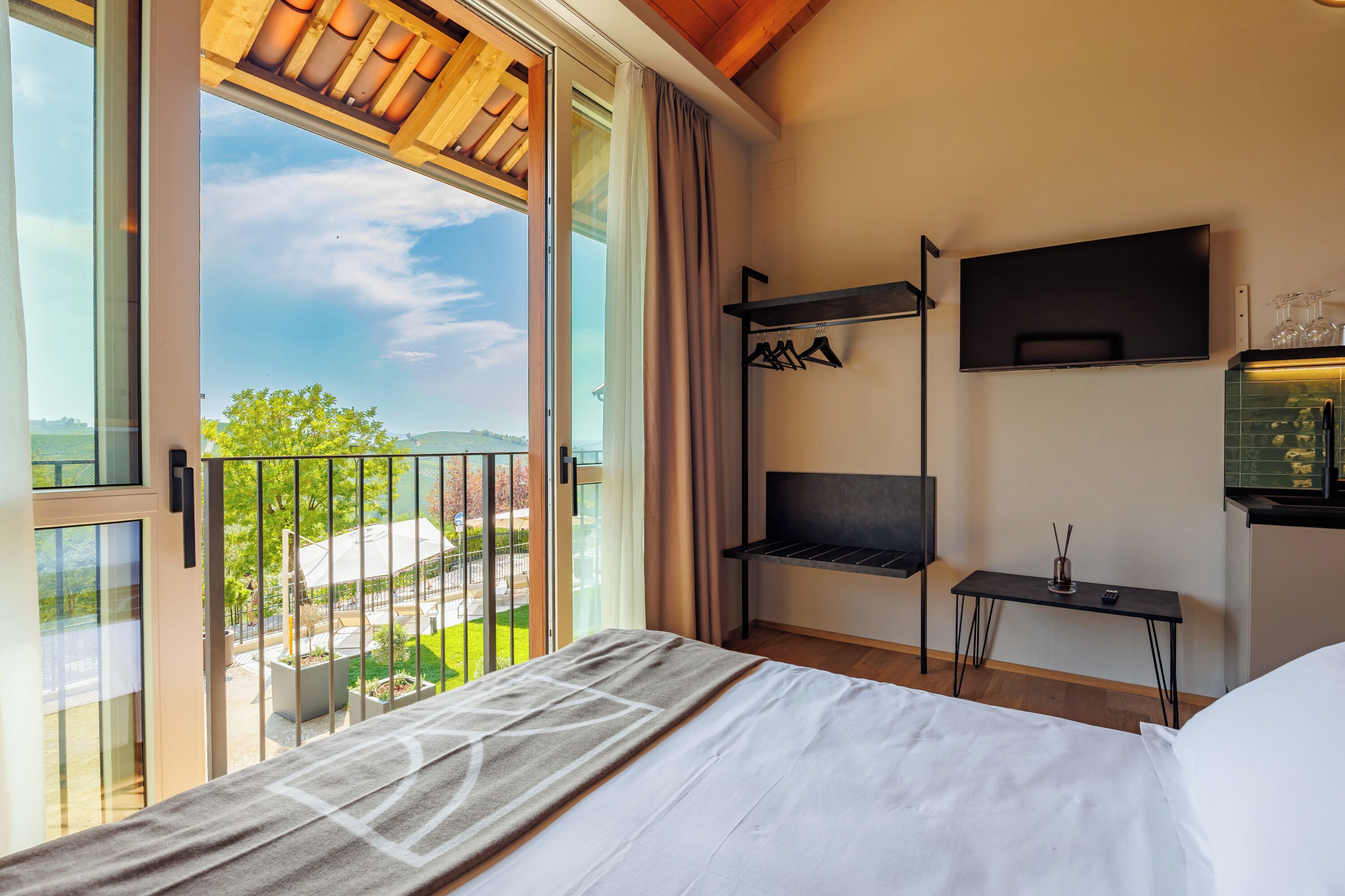 A bedroom with a bed, a TV, a shelf with wine glasses, and a view of a green landscape.