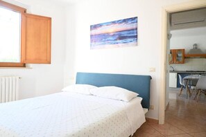 A bedroom with a bed, a painting of a sunset, a window, and a view into the kitchen. - Casa Zefiro – Private Entrance with Courtyard, 10 min from the Beach (Marzocca)