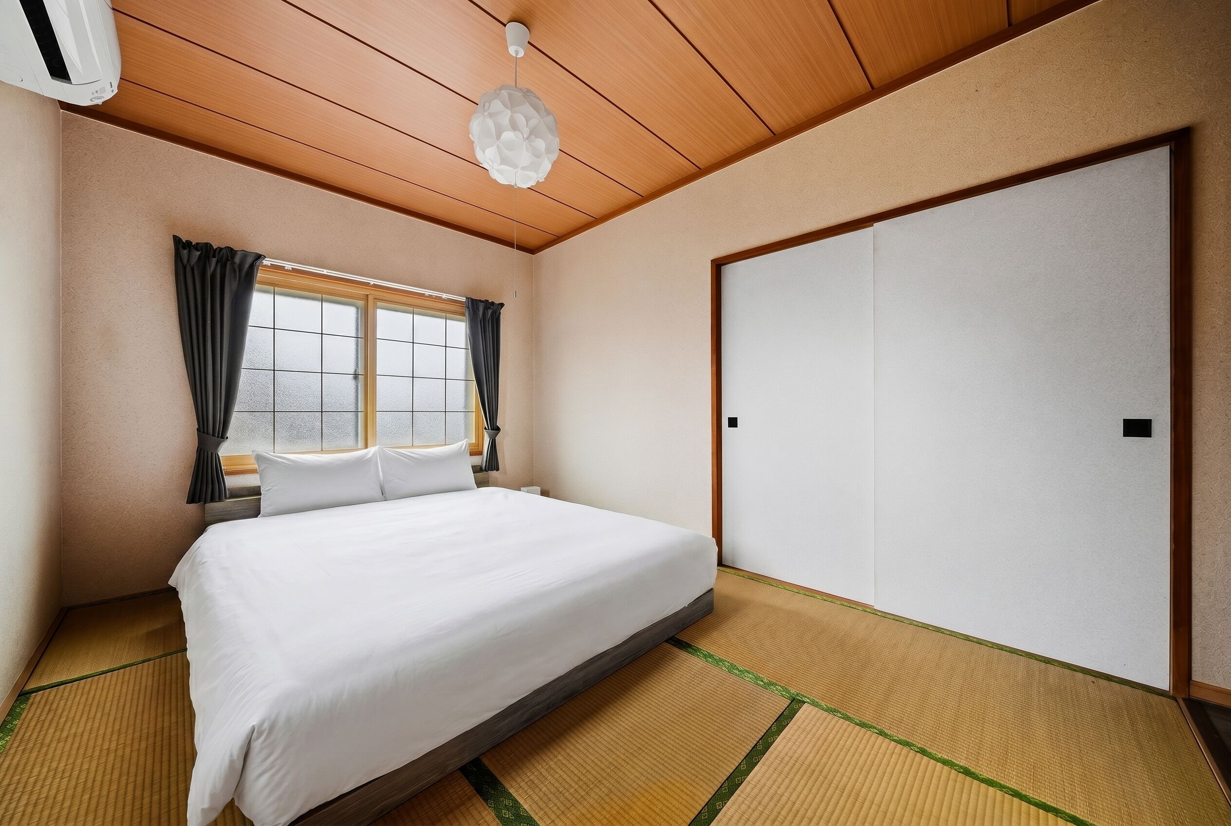 Hotellihuone, jossa on sänky, ikkuna verhoilla, kattotuuletin ja liukuovi.
