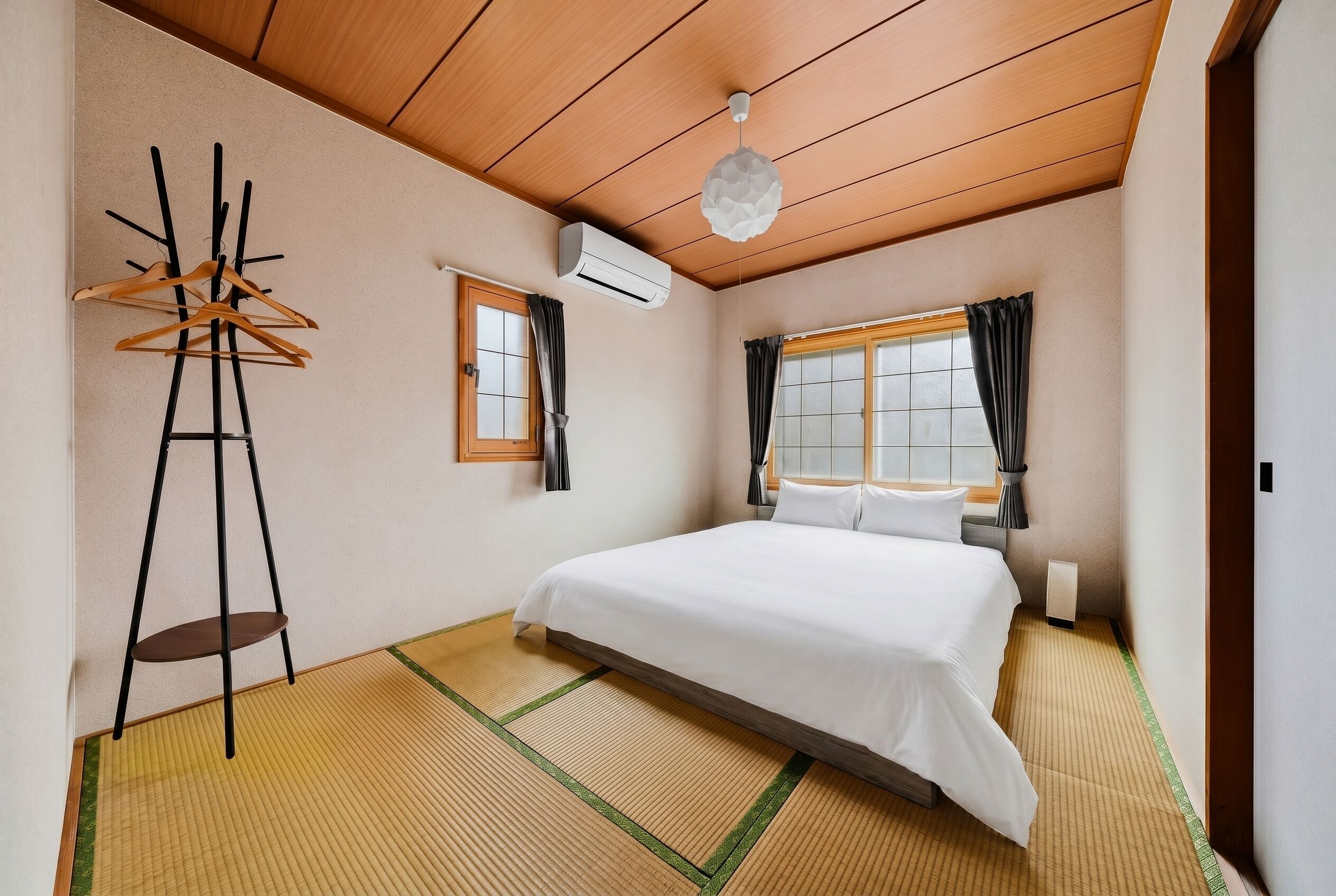 A hotel room with a bed, a hanging coat rack, a window with curtains, and an air conditioning unit.