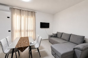 Un salon moderne comprenant une table à manger, deux chaises, un canapé gris, un téléviseur fixé au mur et une fenêtre agrémentée de rideaux légers.