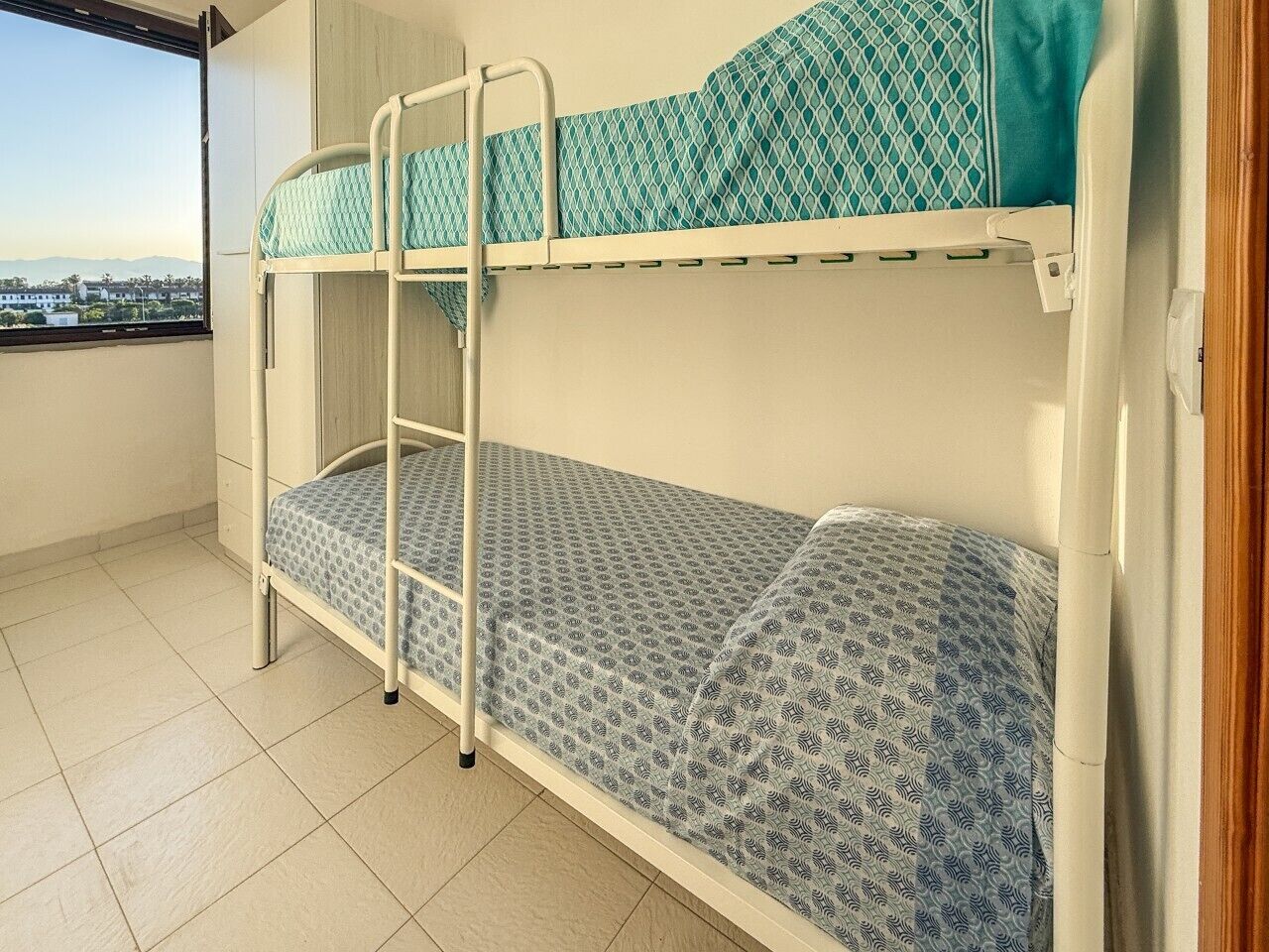 Een stapelbed met beddengoed met een patroon, een trapje en een raam met uitzicht op gebouwen.