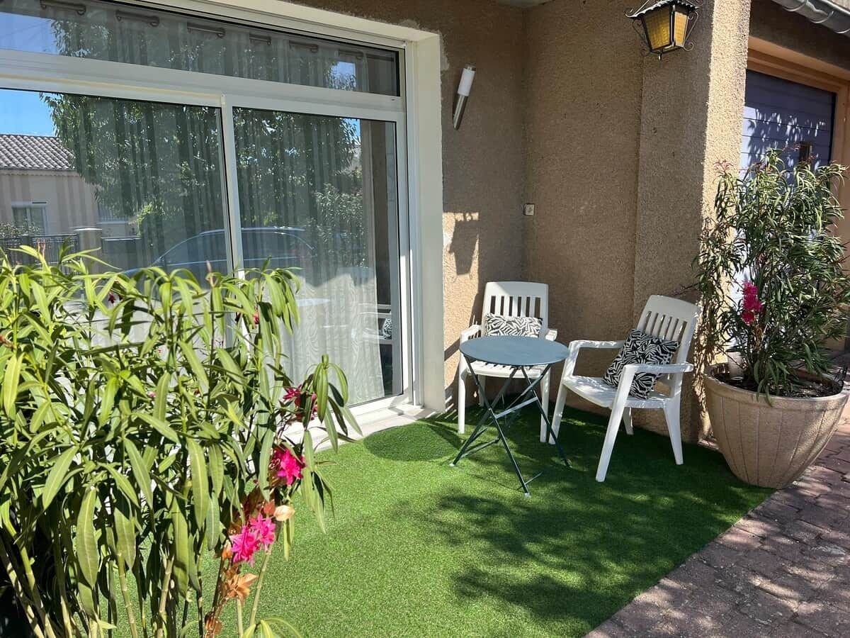 Un patio con césped artificial, dos sillas blancas, una mesita y una planta en maceta.