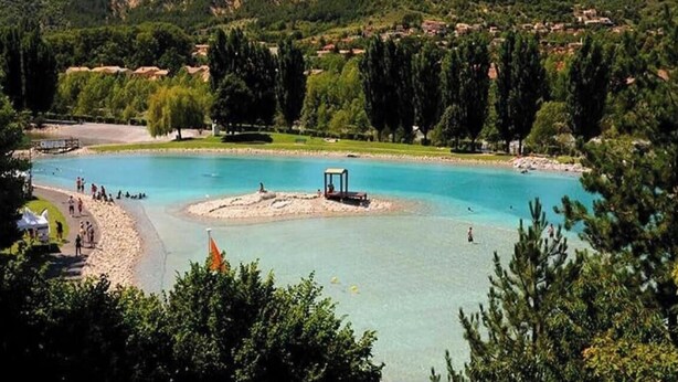 Un lago con una piccola isola, circondato da vegetazione e con edifici sullo sfondo.