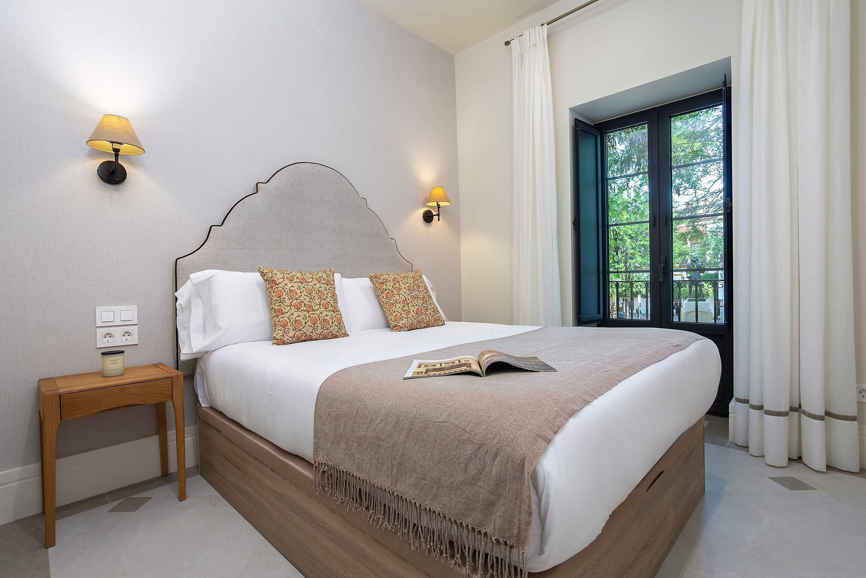 Una cama bien hecha con cabecera, lámparas de noche, una mesita de madera y una ventana con cortinas.