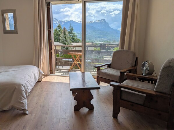 Une pièce comprenant un lit, une table en bois et une chaise. Il y a un balcon d’où l’on peut admirer la vue sur les montagnes et les arbres.