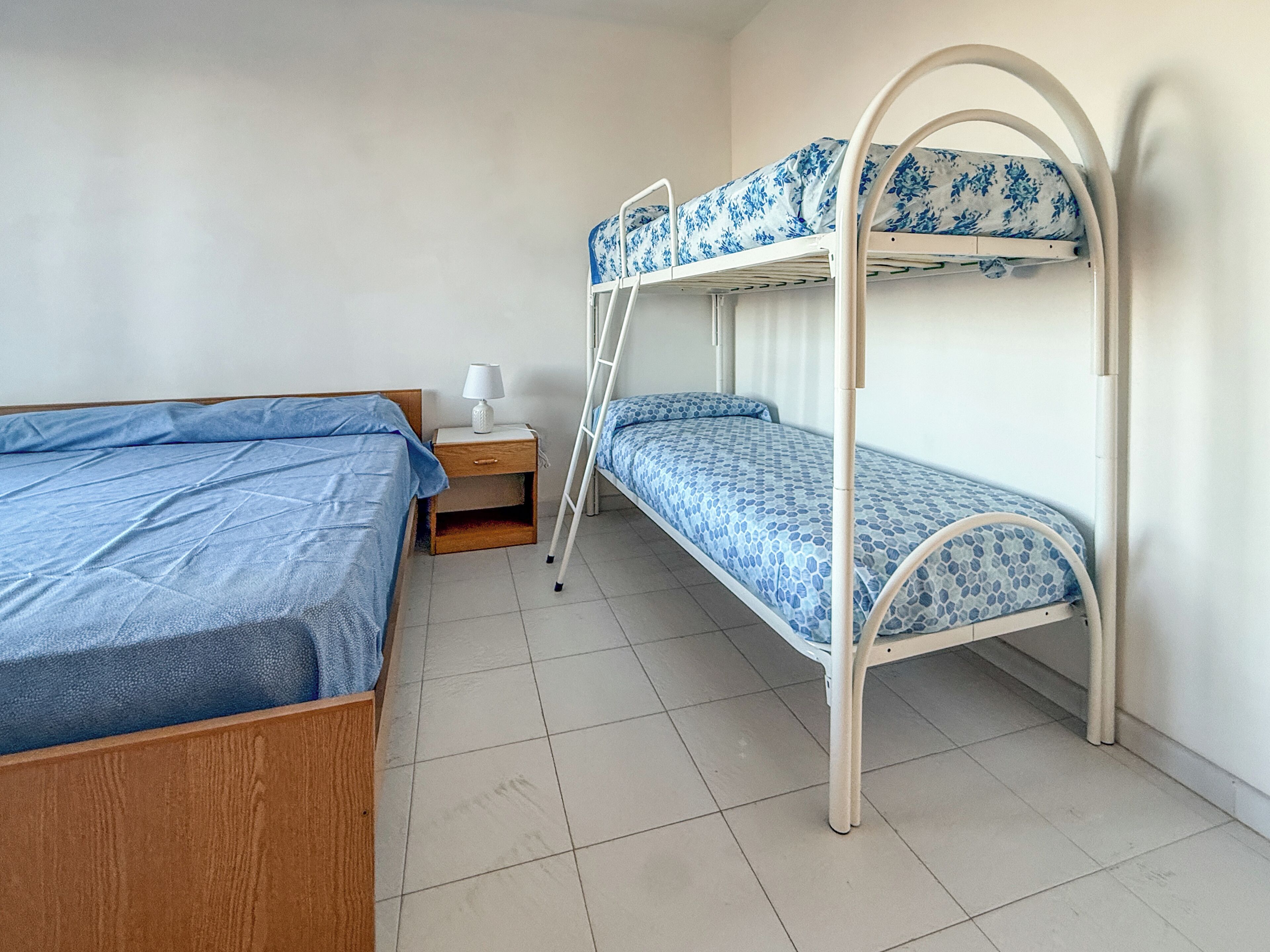A room with two beds, one wooden and one metal bunk, a nightstand with a lamp, and a tiled floor.