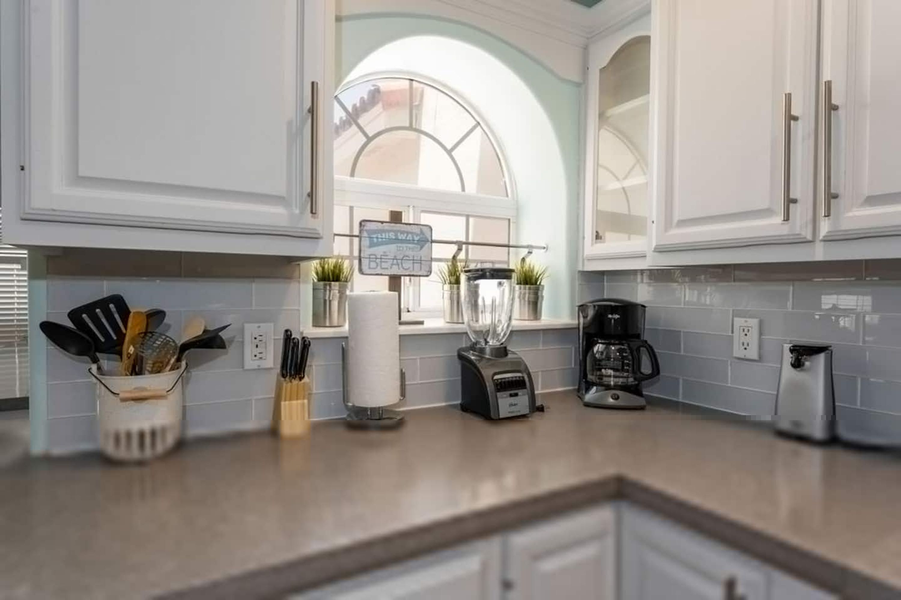 A kitchen with white cabinets, a window with a view, a blender, a coffee maker, and a sign.