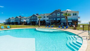 A swimming pool with blue chairs, a palm tree, and a row of multi-story buildings with balconies. - Private Path to Whitecap Beach Pool 5 Stars (Corpus Christi)