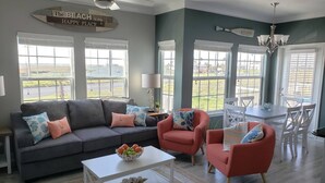 A living room with a grey sofa, red armchairs, a white coffee table, and a dining table with chairs. There are large windows with blinds, a ceiling fan, and a sign on the wall. - Private Path to Whitecap Beach Pool 5 Stars (Corpus Christi)