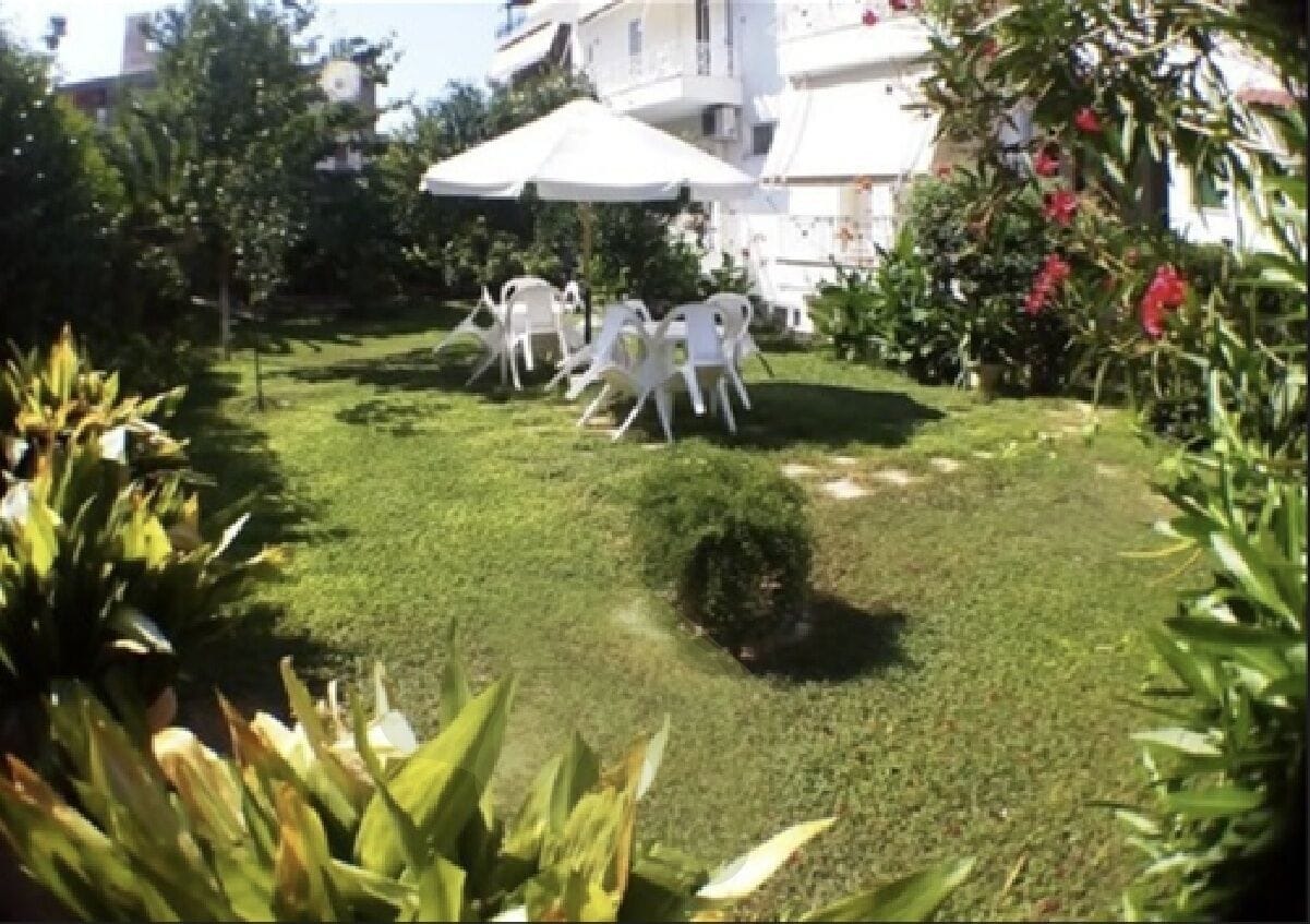 Un jardín cuidado con sillas y sombrillas blancas, rodeado de follaje y plantas con flores.