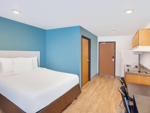 A hotel room with a bed, a desk, a chair, a kitchenette, and a door. - Woodspring Suites Southington (Southington)